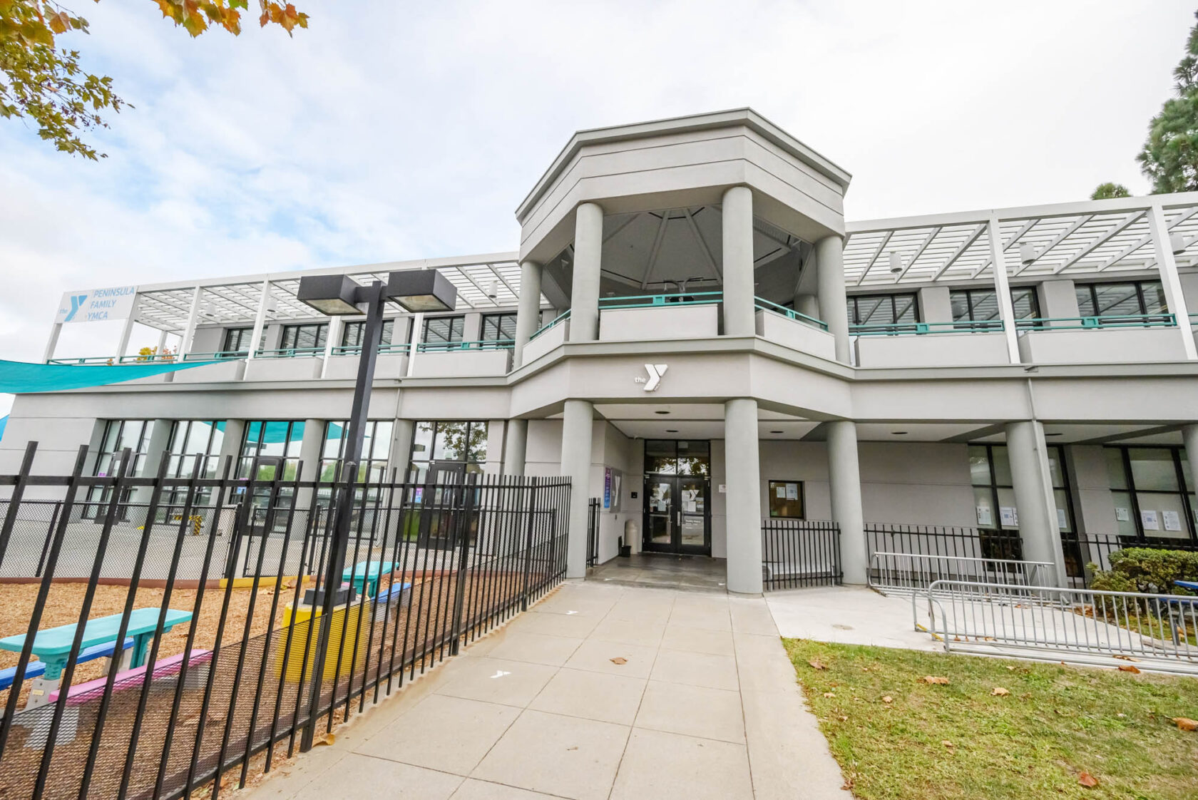 A two-story grey building with a covered porch entrance and Y logo above the door. The front area has a fenced playground with colorful benches. Trees frame the left side and a cloudy sky overhead.
