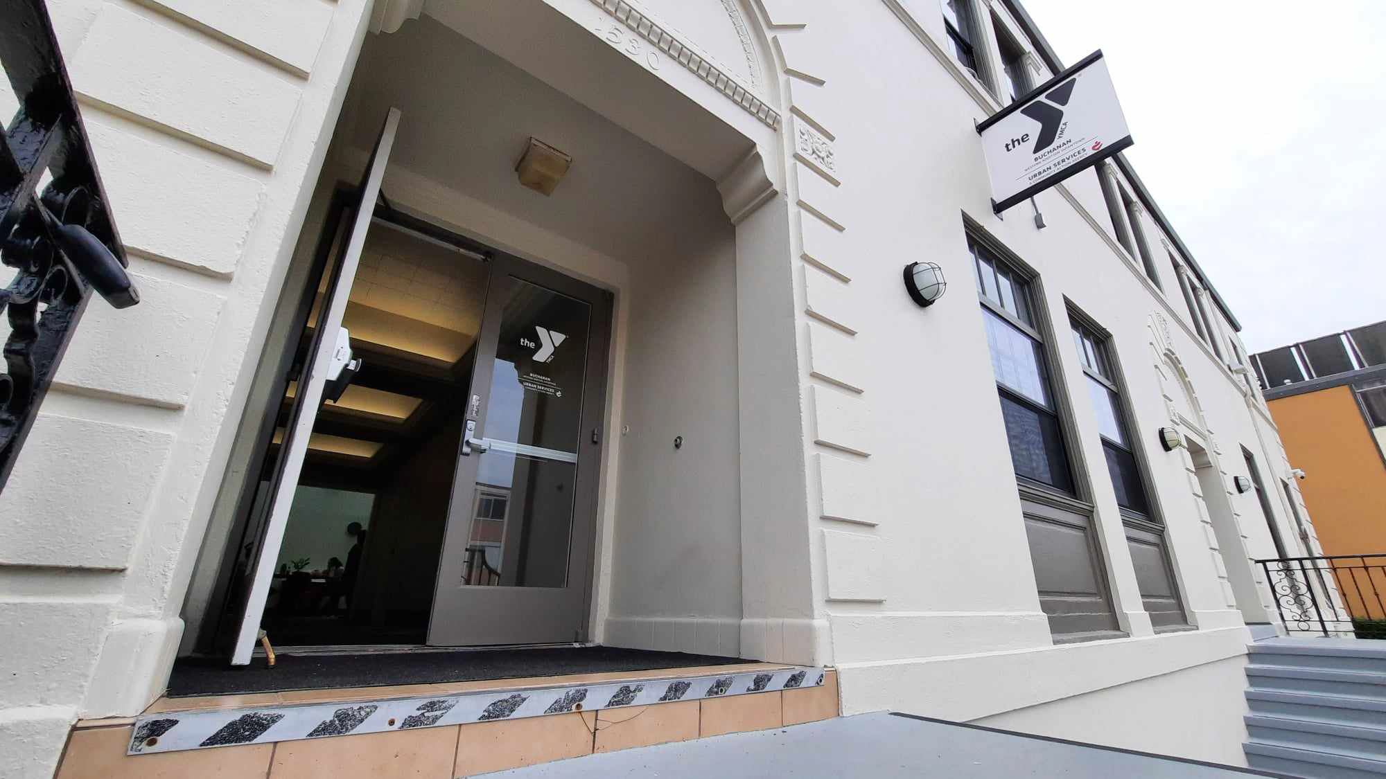 Entrance of a building with an open door, displaying a sign above that reads “YWCA” and a logo. The image shows steps leading up to the doorway and architectural details of the exterior.