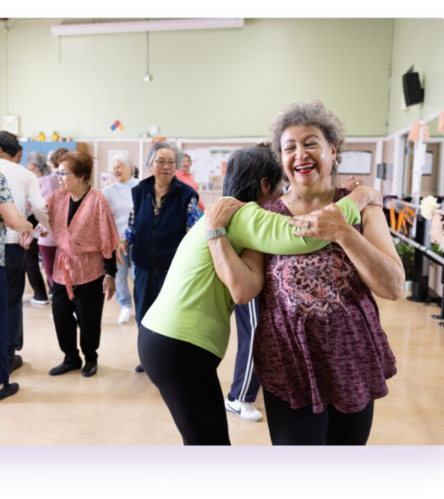 Older adults participate in a group dance or exercise class at the YMCA, with some smiling and embracing while others interact in the background.