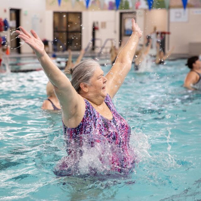 An older woman in a swimsuit raises her arms and splashes water while participating in a group water exercise class at the Peninsula Family YMCA’s indoor swimming pool.