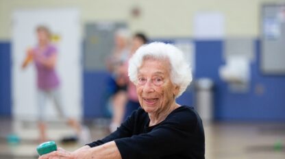 An older woman in black exercise clothes lifts a green dumbbell during a community fitness class at Presidio Community YMCA, smiling at the camera, with other participants working out in the background at this San Francisco gym.