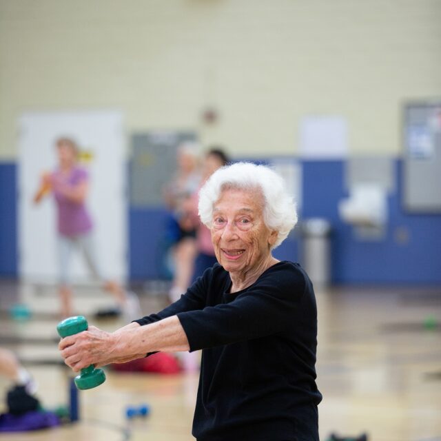 An older woman in black exercise clothes lifts a green dumbbell during a community fitness class at Presidio Community YMCA, smiling at the camera, with other participants working out in the background at this San Francisco gym.
