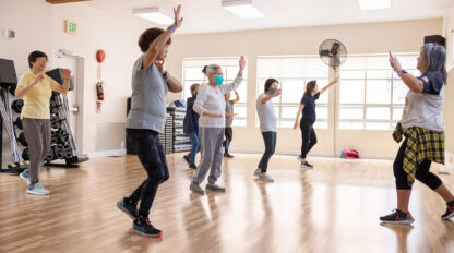 A group of people in casual sportswear practice a dance routine in a brightly lit studio with wooden floors.