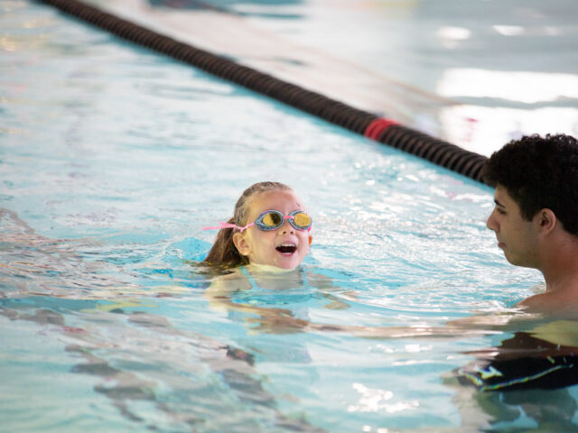 A young child wearing goggles and a swim cap is swimming in a pool while being watched by an adult instructor.