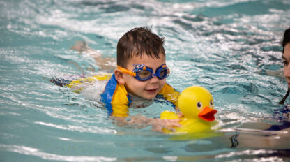 A child in a yellow and blue swimsuit is swimming in a pool while holding a yellow rubber duck.