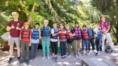 A group of children and two adults in life vests stand and pose outdoors in a forested area, some giving thumbs up. They are under trees with bright sunlight filtering through the leaves.