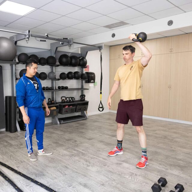 Two men are in a gym; one man in a blue tracksuit observes while the other man lifts a kettlebell overhead with his right hand.