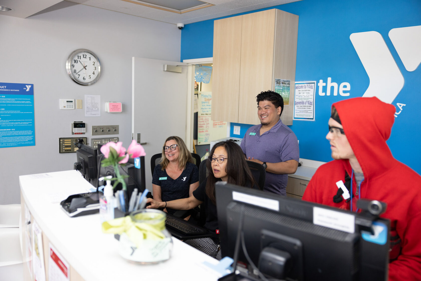 Four people, including one in a red hoodie, are seated and standing around a computer desk in an office with the 'Y' logo on the wall. A wall clock and various signs are visible in the background.