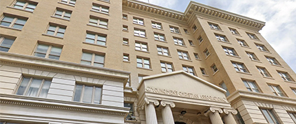 A multi-story beige building with numerous windows. The entrance features a portico with "Commonwealth Building" inscribed on it. Pillars support the portico, and the sky is visible in the background.