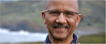A man wearing glasses and a plaid shirt smiles while standing outdoors with a blurred natural landscape in the background.