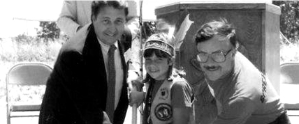 Three people posing for a photo outdoors, one in a suit, one in a feathered headband and sash, and one in a casual shirt with glasses, all smiling. Chairs and a podium are visible in the background.