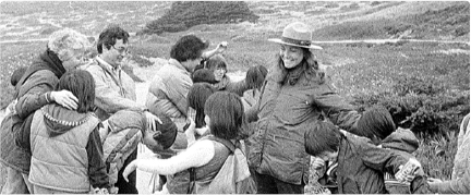 A group of people, including children and adults, stand outdoors in a mountainous area. They seem to be exploring or learning about the environment, as one adult in a hat leads a discussion.