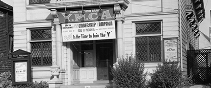 Black and white photo of a YMCA building with a banner advertising a membership campaign and the slogan "Now is the Time to Join the Y" hung above the entrance.