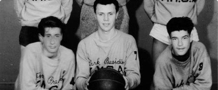 A black-and-white photo of three young men in matching jerseys. The center person is holding a basketball, while the two others sit on either side. Two more people are partially visible standing behind them.