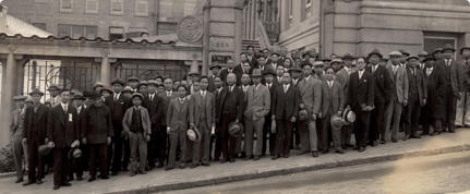 A large group of men in suits and hats stand in front of a building, posing for a formal photo.