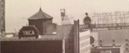 Black and white photo of urban buildings showing signs for YMCA rooms and Sherwin-Williams paint.