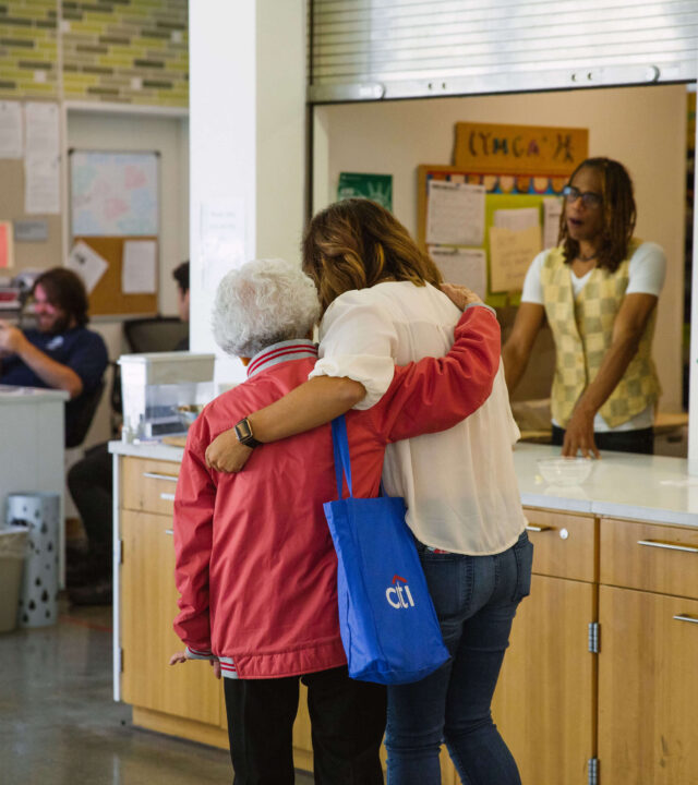 Two women, one elderly and one younger, stand with arms around each other in a community center where others chat and participate in activities like reformer pilates in the background.