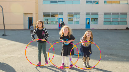 Three elementary aged girls standing in a schoolyard, each holding a hula-hoop and smiling.