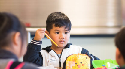 A young boy holding a pencil, seated at a table with a small yellow Pikachu backpack.