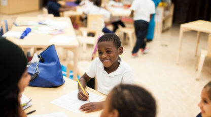 A smiling boy is seated at a table with a pencil and a piece of paper.