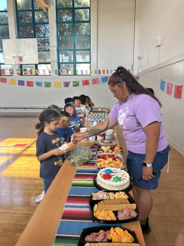 在装饰着彩色横幅的基督教青年会体育馆里,一位女士正在为孩子们分发食物,桌上摆放着各种零食、水果和蛋糕。