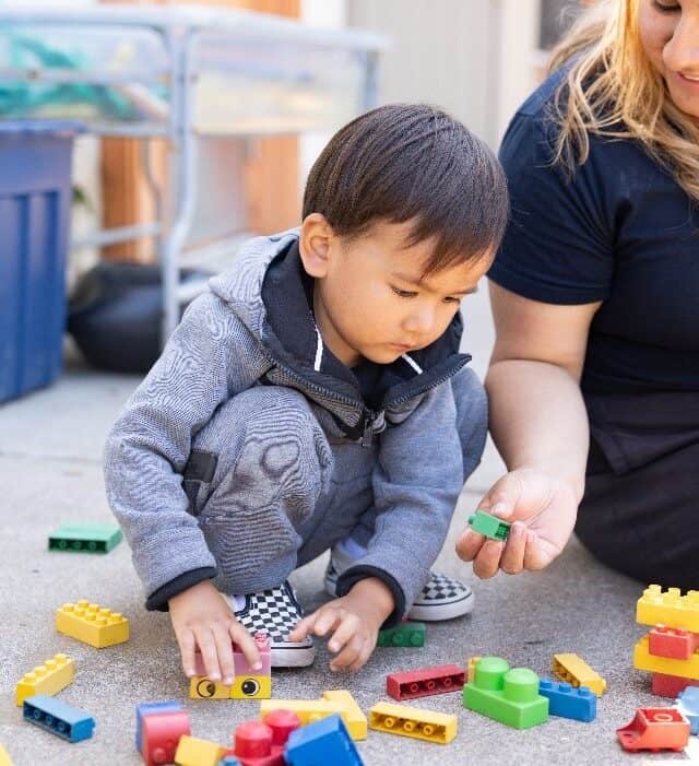 A young child and an adult sit on the ground outside, playing with colorful plastic building blocks after their swim lessons at the YMCA.