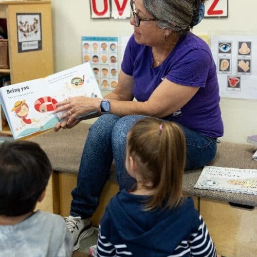 A teacher reads a picture book to two young children in a classroom with alphabet and educational posters on the wall, similar to learning spaces at the YMCA, where kids also enjoy activities like swimming pool fun and beginner reformer pilates.