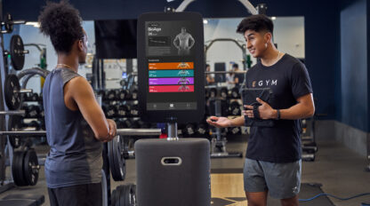 Two people in a gym stand near a touchscreen fitness device displaying workout details; one explains the information to the other. Various gym equipment is visible in the background.