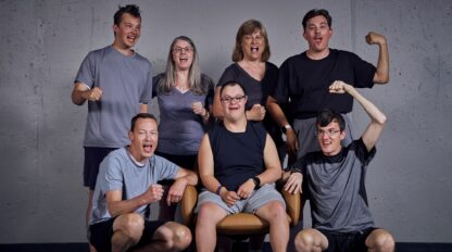 Seven people pose together, with two seated in front and five standing behind them, all smiling and making energetic gestures in a casual indoor setting.