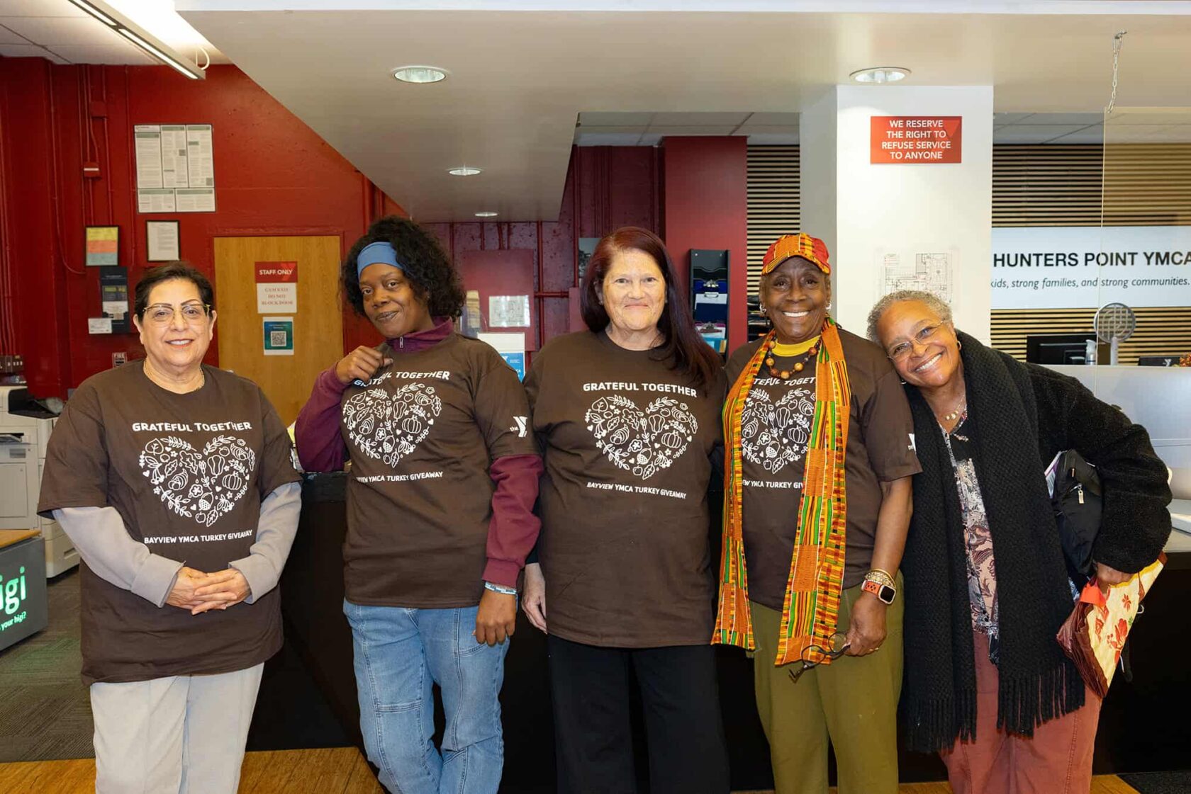 Five women stand side by side indoors, four wearing matching "Grateful Together" shirts, smiling at the camera in a community center setting.