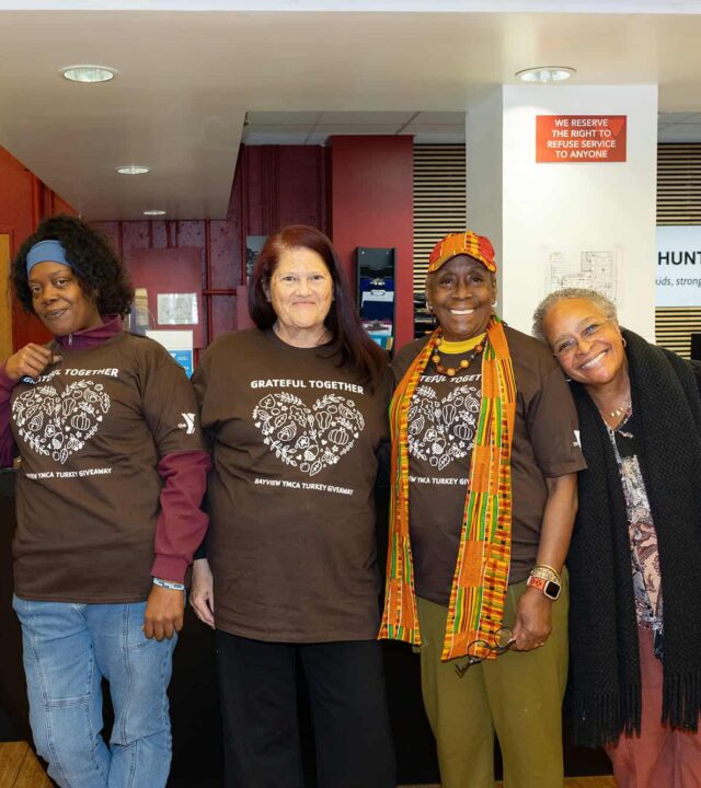 Five women stand side by side indoors, four wearing matching "Grateful Together" shirts, smiling at the camera in a community center setting.