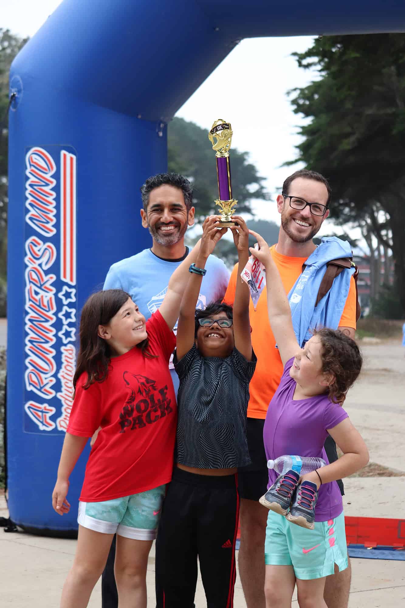 Two adult males and three youth standing at the finish line of a race while holding up a trophy.