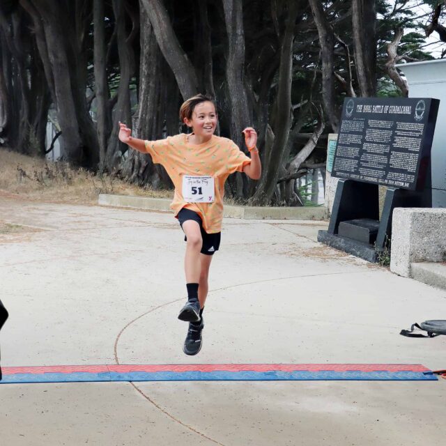 Youth male jumping in the air with his arms raised as he crosses the finish line.