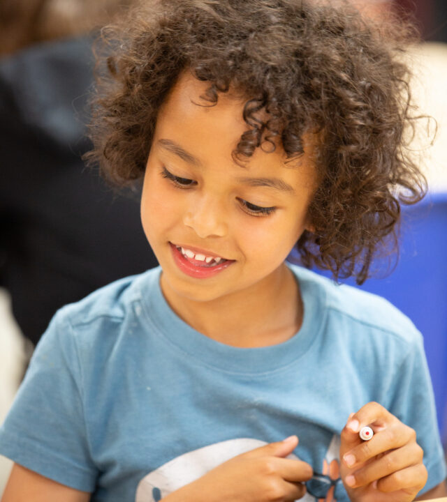 A smiling young child seated at a table holding a marker in their hand.