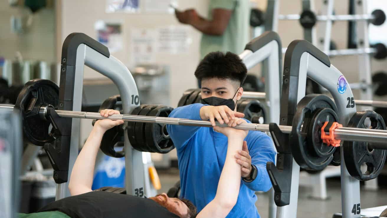 One adult male showing another adult male a weight lifting technique.