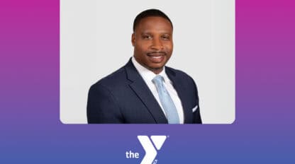 Derick Brown, dressed in a dark suit and light tie, poses for a formal portrait against a white background, with the YMCA of Greater San Francisco “the Y” logo below on a pink-to-blue gradient backdrop highlighting Vision 2030.