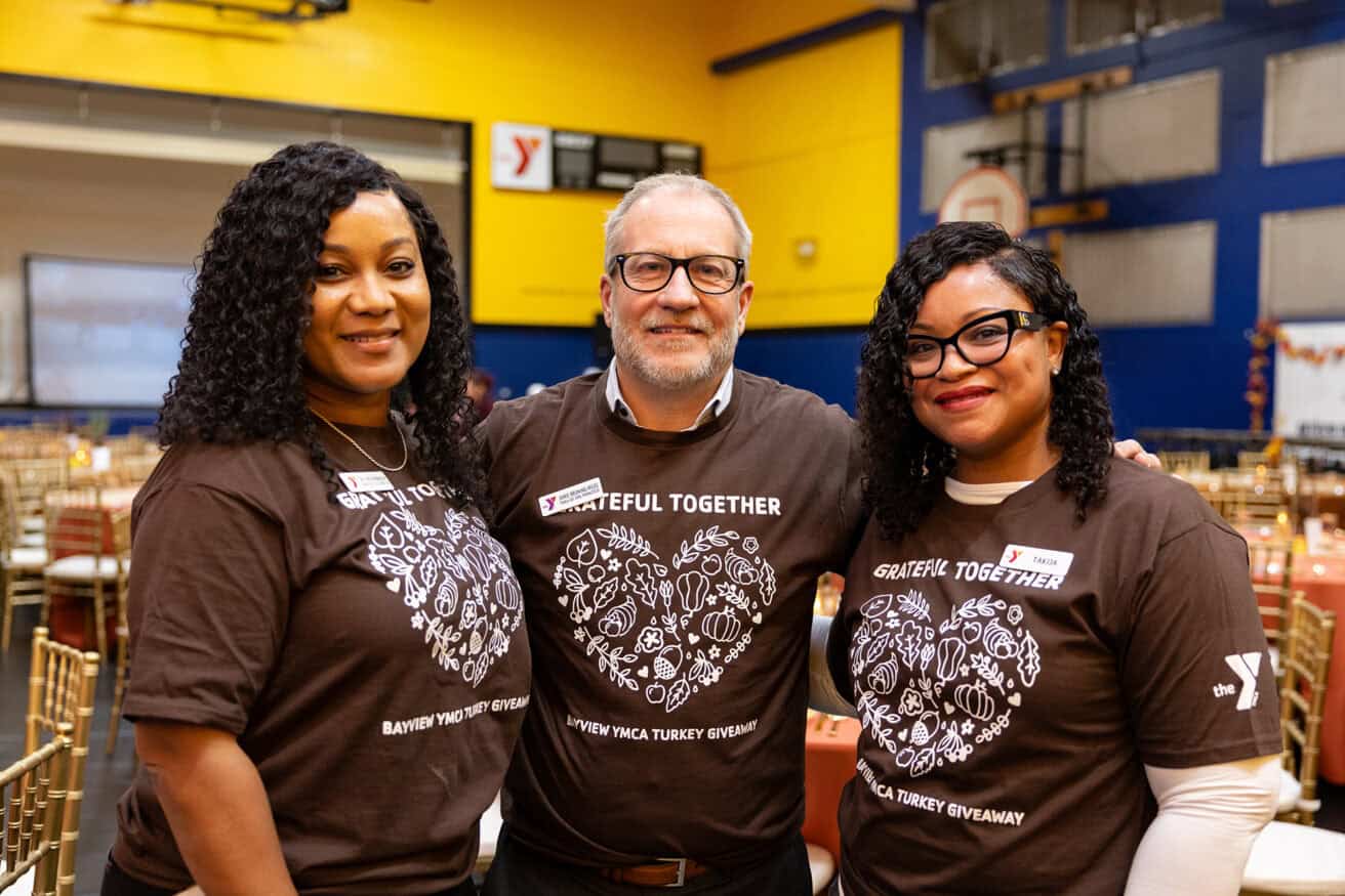 Three community leaders stand side by side in a decorated gymnasium, wearing matching "Grateful Together" shirts and smiling at the camera, embodying the spirit of strong communities.