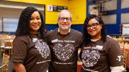 Three community leaders stand side by side in a decorated gymnasium, wearing matching "Grateful Together" shirts and smiling at the camera, embodying the spirit of strong communities.