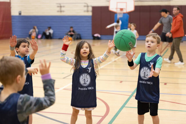 Four young children in Presidio YMCA jerseys stand in a gym, reaching out as a green basketball is passed to them during a Youth Sports Programs activity, celebrating teamwork within the Presidio Community.