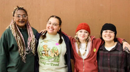 Four people stand side by side in front of a brown wall at Camp Jones Gulch, smiling at the camera with their arms around each other during a YMCA Wellness Weekend.