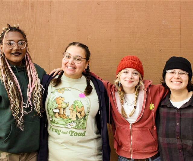 Four people stand side by side in front of a brown wall at Camp Jones Gulch, smiling at the camera with their arms around each other during a YMCA Wellness Weekend.
