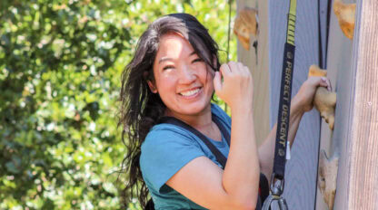 A woman wearing a harness smiles while climbing a rock wall outdoors at Camp Jones Gulch, with green trees in the background, enjoying the spirit of a YMCA Wellness Weekend.