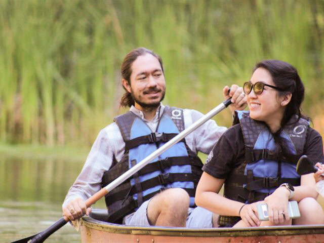 Two people wearing life jackets and sunglasses are sitting in a canoe on a calm body of water at Camp Jones Gulch, holding paddles with tall green reeds in the background during the YMCA Wellness Weekend.