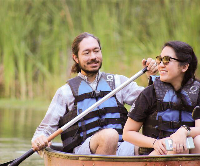 Two people wearing life jackets and sunglasses are sitting in a canoe on a calm body of water at Camp Jones Gulch, holding paddles with tall green reeds in the background during the YMCA Wellness Weekend.