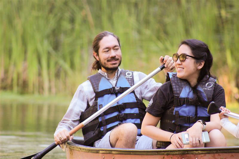 Two people wearing life jackets and sunglasses are sitting in a canoe on a calm body of water at Camp Jones Gulch, holding paddles with tall green reeds in the background during the YMCA Wellness Weekend.