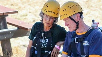Two older adults wearing helmets and harnesses sit next to each other outdoors at Camp Jones Gulch, looking down, with a wooden picnic table in the background.
