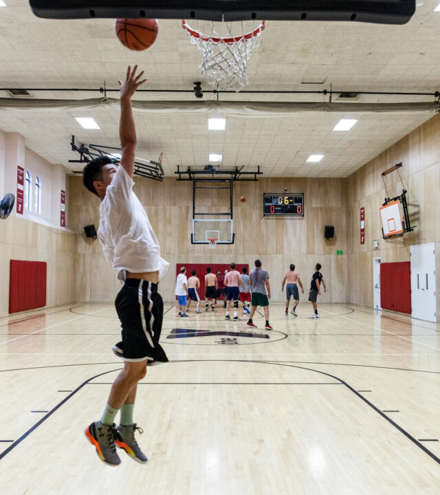 Adult male jumping in the air playing basketball in a gym.