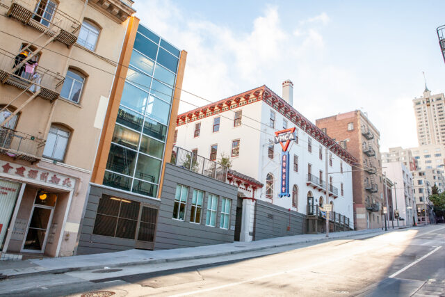 A street view of buildings, including the YMCA of Greater San Francisco Executive Leadership and Board of Directors' facility, features a prominent sign, modern glass structure, and neighboring apartments on a sloped urban street.