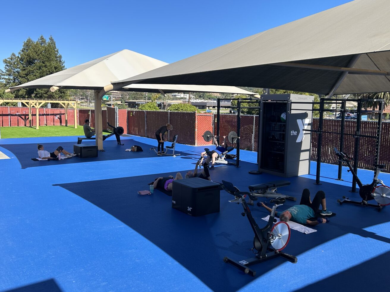 People exercise on mats and stationary bikes under shade structures in the updated gym workout space at Marin YMCA, with fencing and trees in the background on a sunny day.
