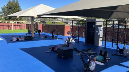 People exercise on mats and stationary bikes under shade structures in the updated gym workout space at Marin YMCA, with fencing and trees in the background on a sunny day.